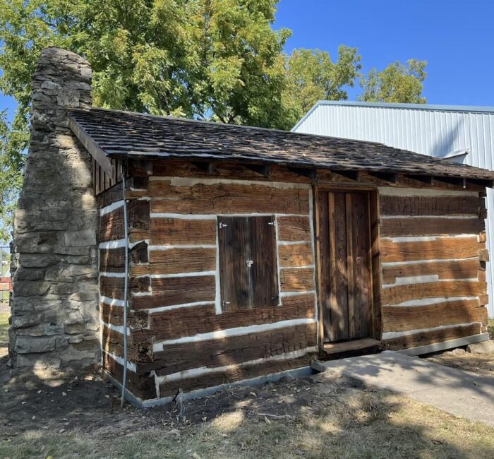Log Cabin Festival Small log cabin with a stone chimney, dark wood logs, white chinking, and a wooden door