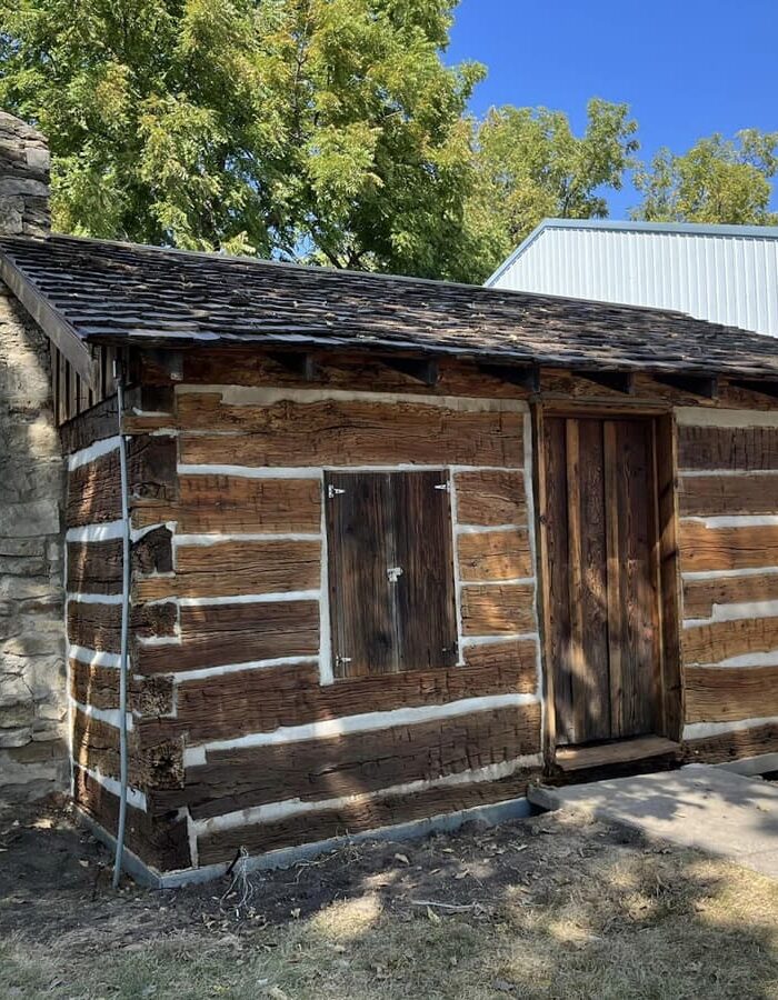 Small log cabin with a stone chimney, dark wood logs, white chinking, and a wooden door