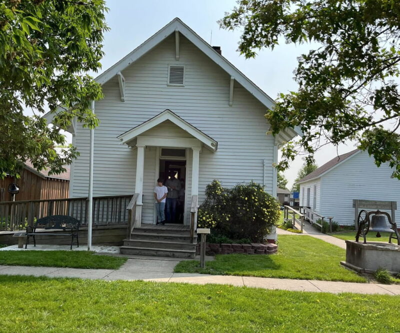 Small white schoolhouse with a gabled roof, wooden siding, a porch, and a large bell on a stone base in the yard