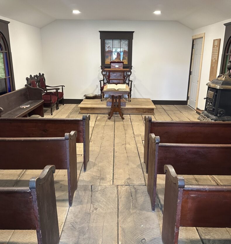 Heritage Village Church Interior of a rustic chapel with wooden pews, wide plank floors, a wood stove, and an open Bible on a pulpit