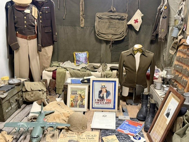 Military memorabilia display with uniforms, field gear, an Uncle Sam poster, and a newspaper announcing Roosevelt's death