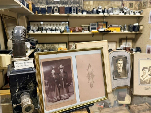 Antique slide projector and framed vintage portraits in front of shelves filled with old cameras