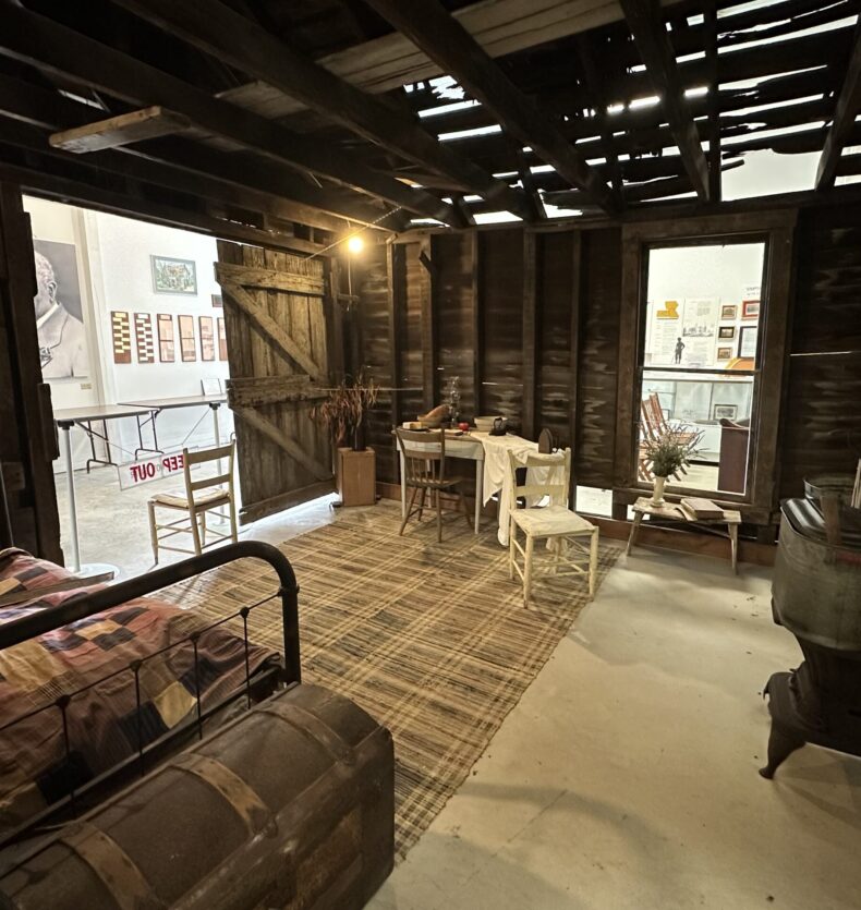 inside Carver house at Warren County Historical Society Museum exhibit of a rustic cabin interior with a bed, trunk, small table, and wood-burning stove