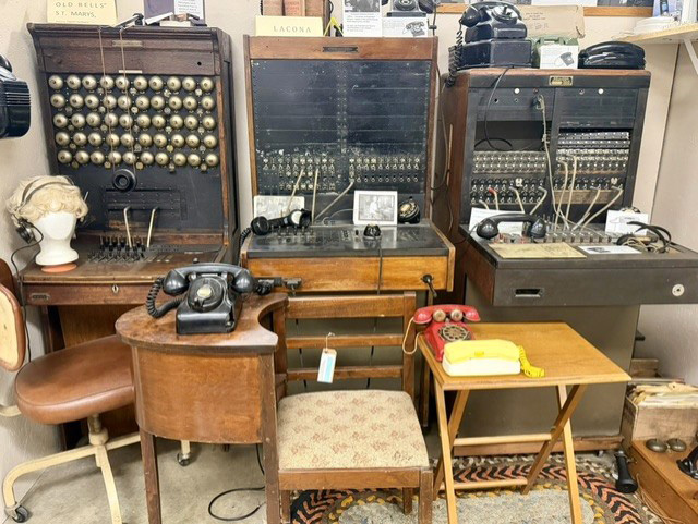 Vintage wooden telephone switchboards with patch cables and bells, surrounded by old rotary and push-button phones