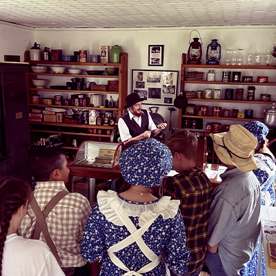 A historical reenactor speaks to a group of visitors in a general store filled with period goods