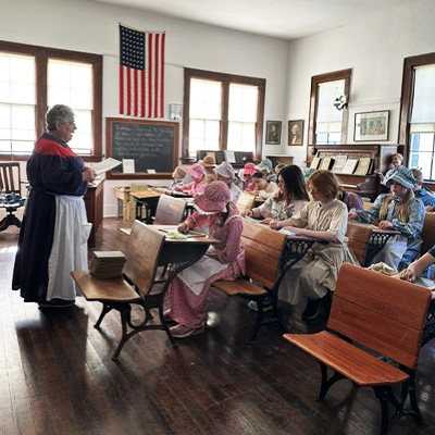 Students in historical dress sit at wooden desks while a teacher reads in a 19th-century style classroom