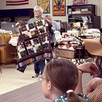 An older woman holds up a multi-colored patchwork quilt for a small audience in a workshop setting