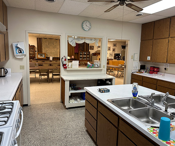 Kitchen with a large island and double sink looking into a dining hall with tables and chairs