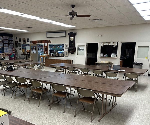 A meeting room with rows of long folding tables and chairs, a ceiling fan, and a grandfather clock